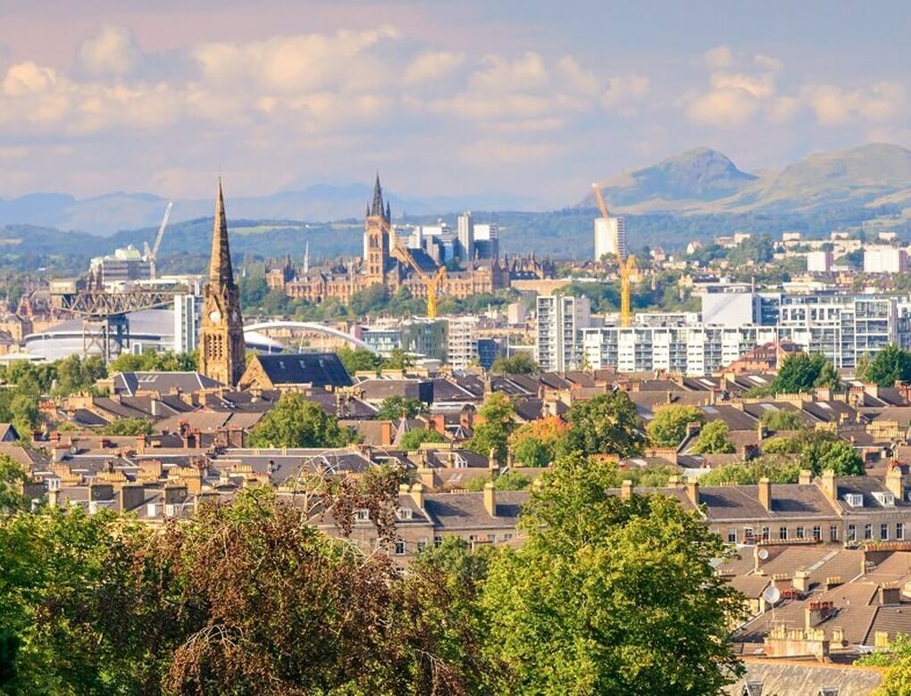 A viewpoint over the city of Glasgow from Queen's Park, taking in the greenery of trees with the city and Scottish landscape in the distance.