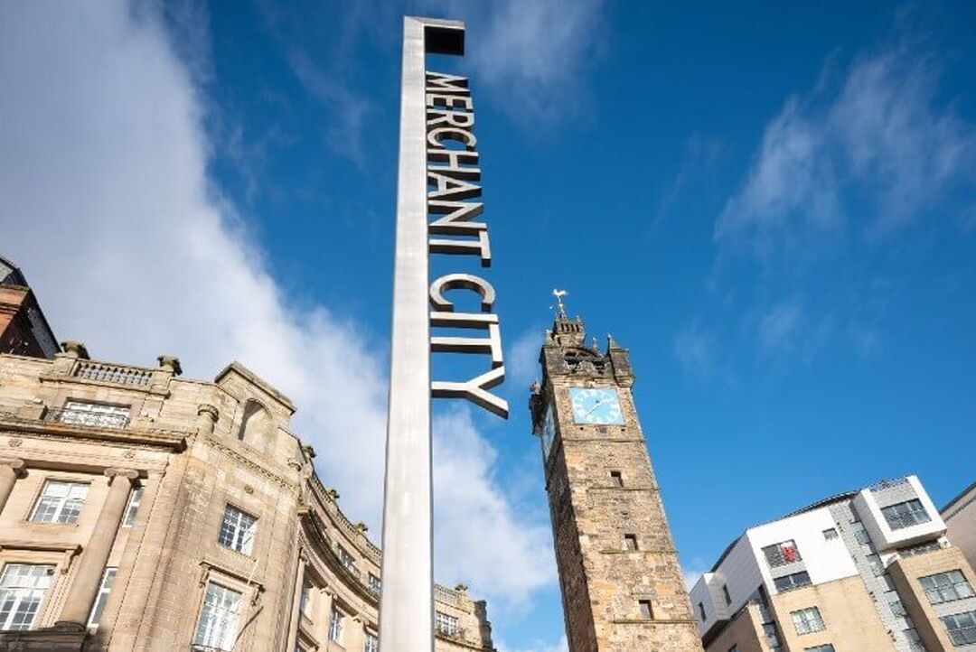 A low angle view of the Merchant City sign, the Tolbooth clock tower can be seen behind the sign.