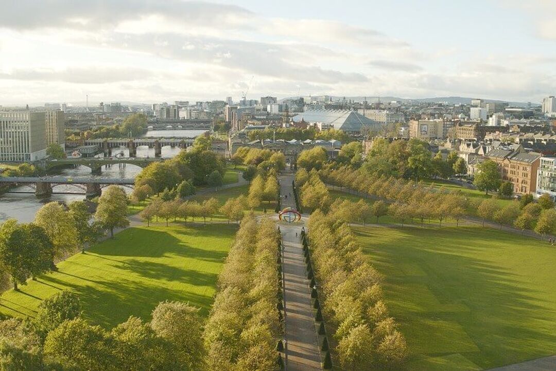 A tree-lined avenue with square and triangle patches of grass to either side. The buildings of the city centre can be seen beyond the greenery, while a river with a number of bridges over it is to the left side.