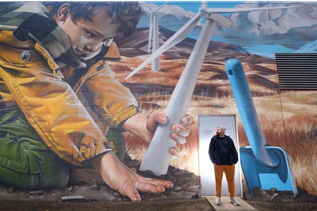 A person is standing in front of one of the mural trail artworks, Generation Green. A young child is planting a windmill in soil, a blue spade is beside them.