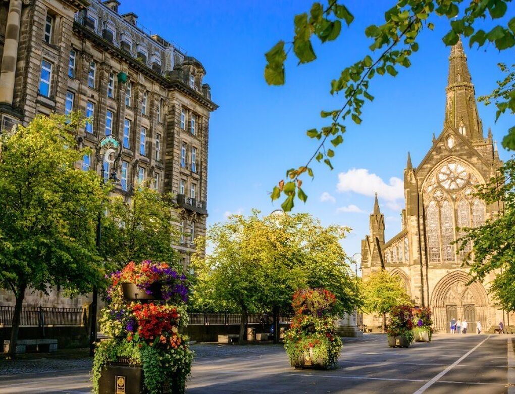 Two historic buildings, including Glasgow Cathedral, surround a cobbled square with many trees and planters