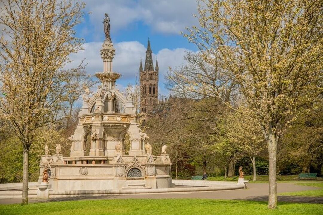 A large fountain in the middle of a park. The gothic tower of Glasgow University can be seen in the background.