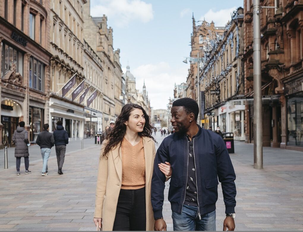 Two people walk down the centre of the pedestrianised Buchanan Street arm-in-arm. On either side of them are grand buildings housing retailers.