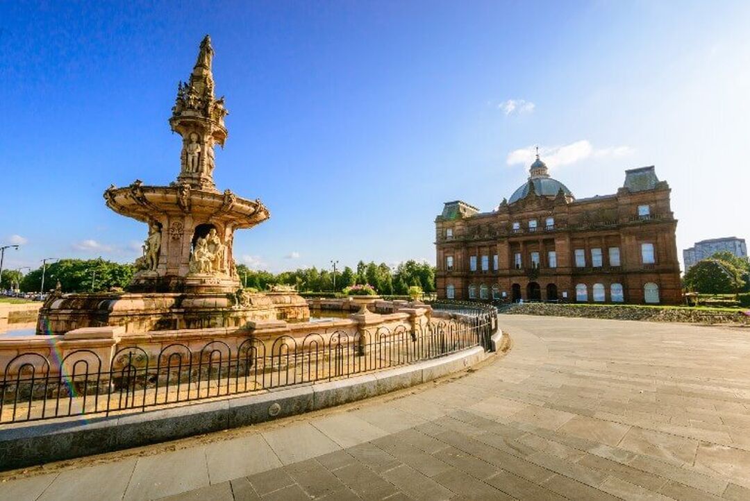 The terracotta Doulton Fountain, which has sculptures of people carved onto it, sits against a blue sky and in front of the red sandstone Victorian People's Palace.