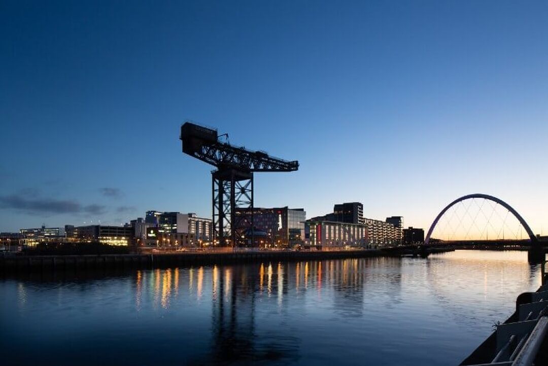 The large Finnieston Crane stands high on the skyline in front of the River Clyde. Lights reflect in the water as the sun sets behind the semi-circle Clyde arc bridge.