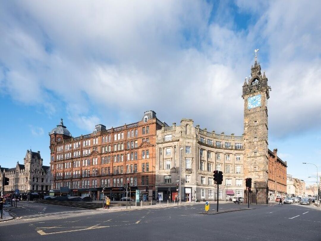 The tall narrow Tolbooth Steeple, which has a clock at the top, sits in the centre of a Glasgow road junction. Behind the steeple stand a row of historic and in use buildings in red and blonde sandstone.