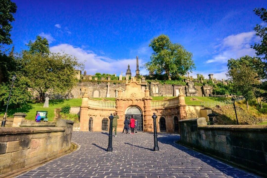 People stand on a cobbled path in front of a stone archway at the entrance to the Glasgow Necropolis. Behind the entrance tombstones and sculptures line the sides of a grassy hill.