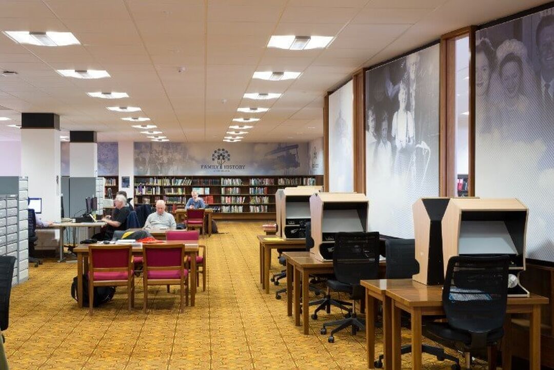 People sit at research desks in front of a wall filled with books and a sign reading 'Family History'. Another wall has large black and white prints of photographs from the past.