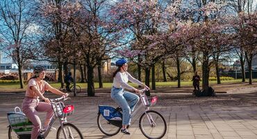 Two people using hired bikes to enjoy cycling around Glasgow Green with cherry blossom trees in the background.