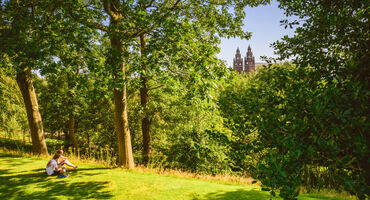green area with trees and person sitting on the ground. Building with tall spires in the background