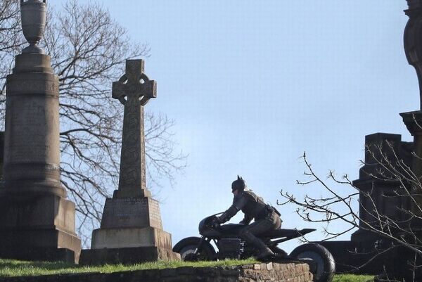 Batman on the Bat Bike drives between monuments in a graveyard.