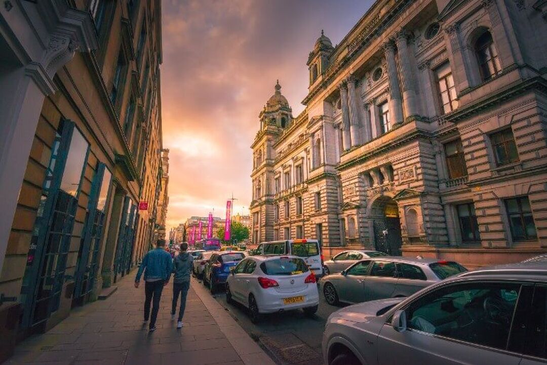 The sun is beginning to set between the buildings of a long street which is lined with grand blonde sandstone buildings.