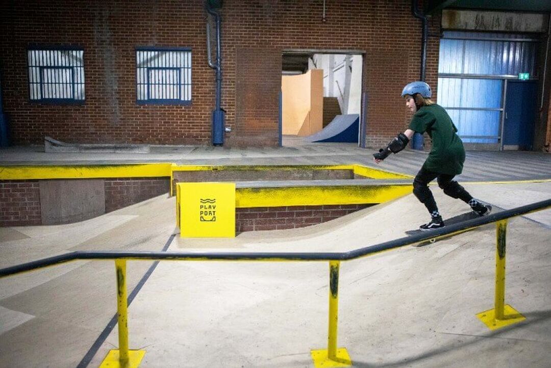A skateboarder goes down a ramp within an indoor skate park that includes rails, ramps and a yellow sign that reads "Play Port".