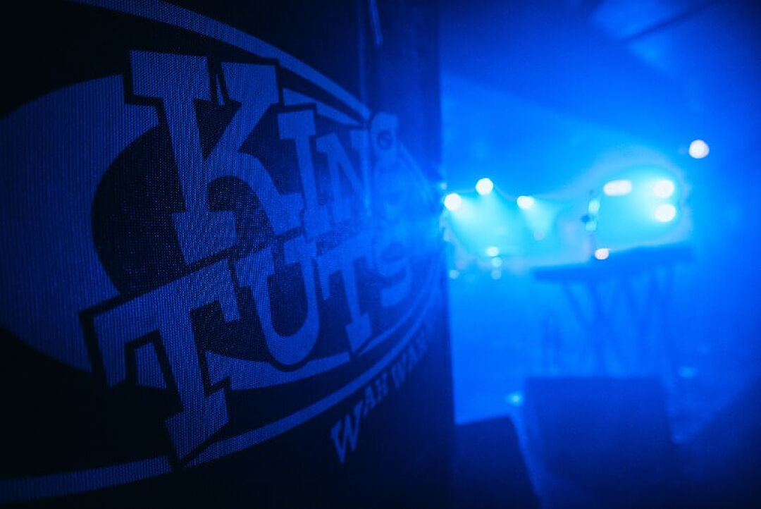 The King Tuts logo is on a wall. Blue light from the stage behind streams across the room.