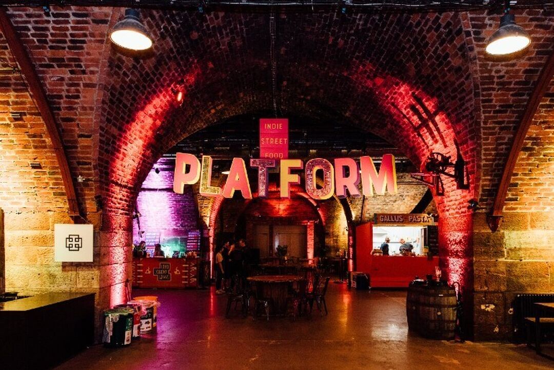Letters spelling out 'Platform' hang beneath a stone archway. The dark space with no natural light is lit up with pink and white lighting.