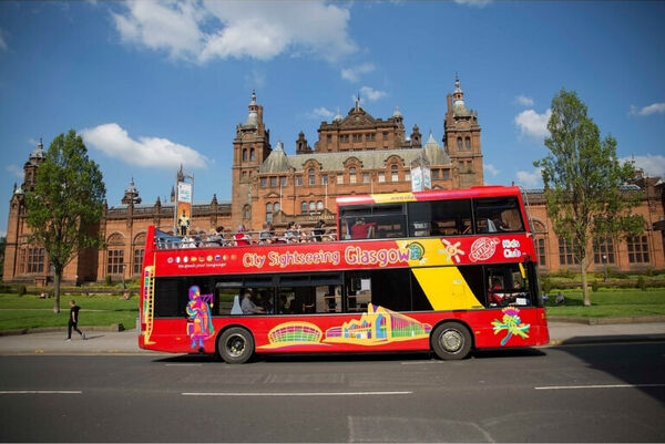 Bright red and yellow open-top bus passing in front of large red sandstone building