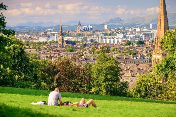 Two people lie on the grass in the sunshine at the top of a park, which has a brilliant view of the city and beyond. The trees open up to reveal a view that includes church steeples, bridges and the Campsie Fells in the distance.