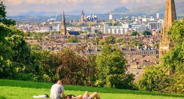 Two people lie on the grass in the sunshine at the top of a park, which has a brilliant view of the city and beyond. The trees open up to reveal a view that includes church steeples, bridges and the Campsie Fells in the distance.