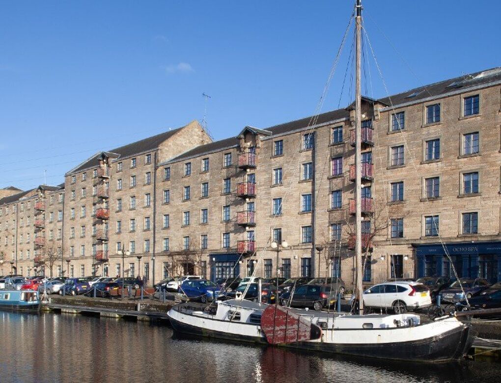 Georgian style buildings line a canal on a sunny day. Boats and barges are berthed in front of the buildings.