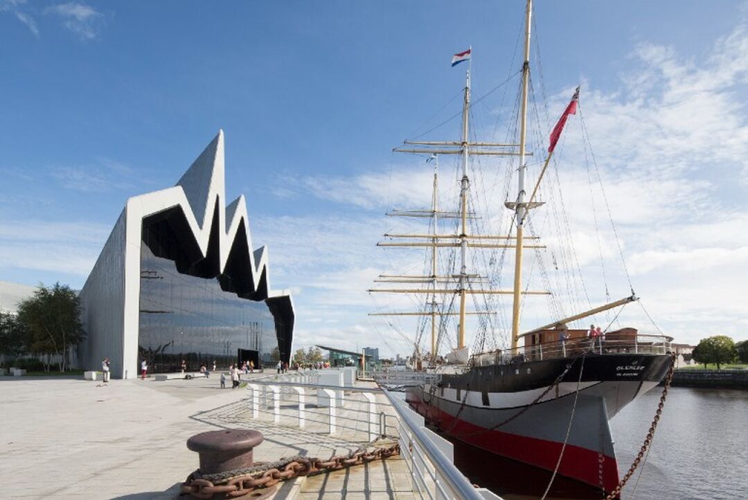 A sail ship is berthed on the river beside a modern building, which is silver with a jagged wave-like roof.