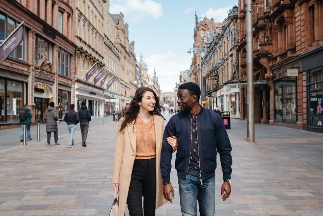 Two people walk down the centre of the pedestrianised Buchanan Street arm-in-arm. On either side of them are grand buildings housing retailers.