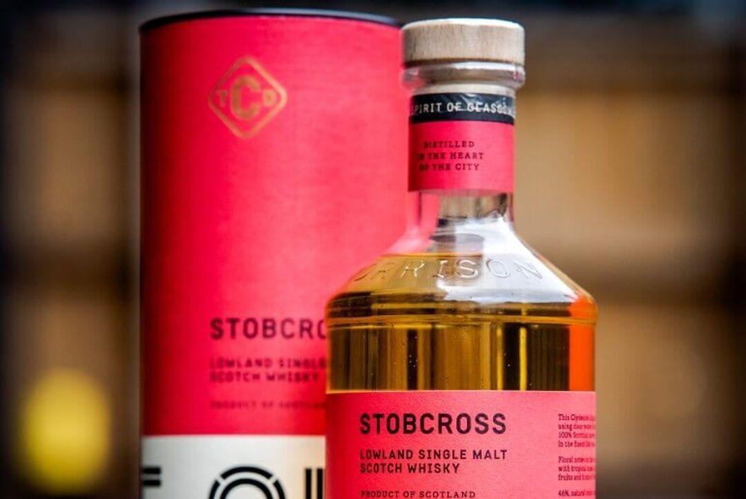 A close-up of a whisky bottle with a red label that reads 'Stobcross' with the red box sitting behind it.