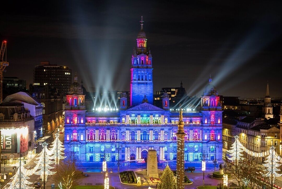 Glasgow City Chambers illuminated in blue and pink lights with a festively decorated George Square beneath.