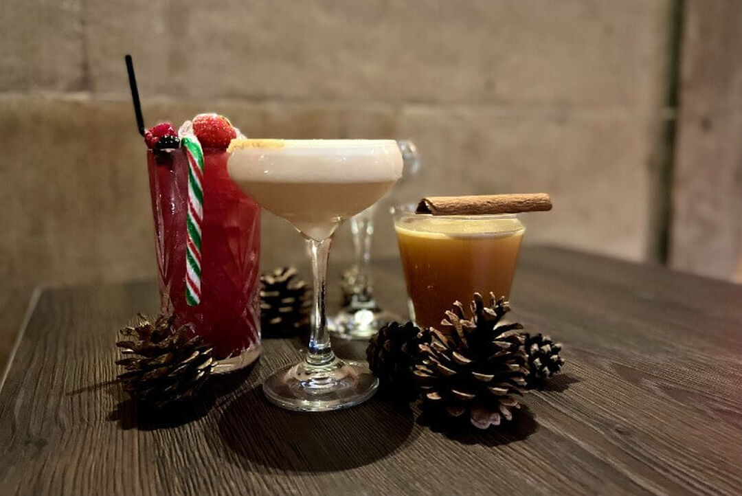 Three cocktails are lined up on a table beside pine cones. A candy cane and cinnamon stick sit on top of the festive drinks.
