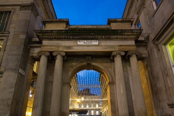 A neo-classical style archway leads towards a canopy of fairy lights. A plaque reading 'Merchant City' sits above the arch.