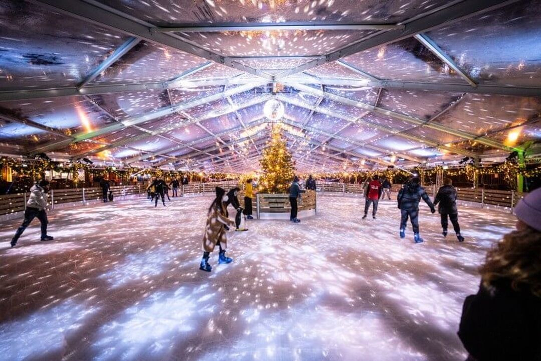 Skaters skate around a circular ice rink which has a Christmas tree in the centre. Fairy lights and a disco ball create sparkles on the ice and the white canopy ceiling.