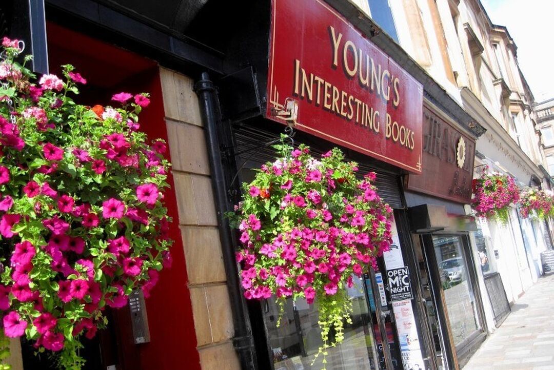Hanging baskets full of pink flowers hang in front of a row of small shops. The signage of the main shop in view says 'Young's Interesting Books'.