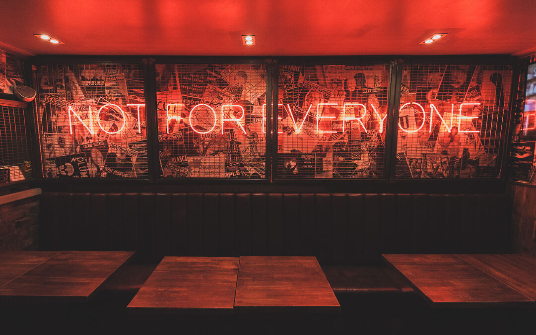 A dark room with dimly lit red lights, the wall has is lit up with red lettering saying "Not For Everyone". Behind the letters, is a collage of photos. Below the wall are a row of tables.