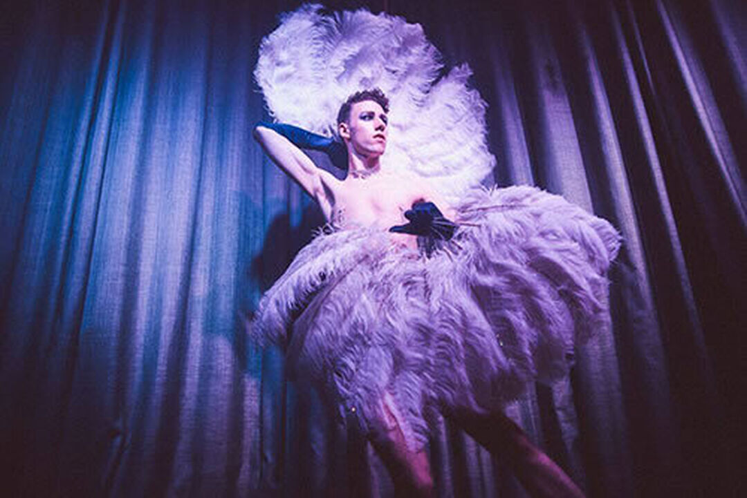 A person who is topless dancing on a stage with two large feather canopies in front of a blue curtain.