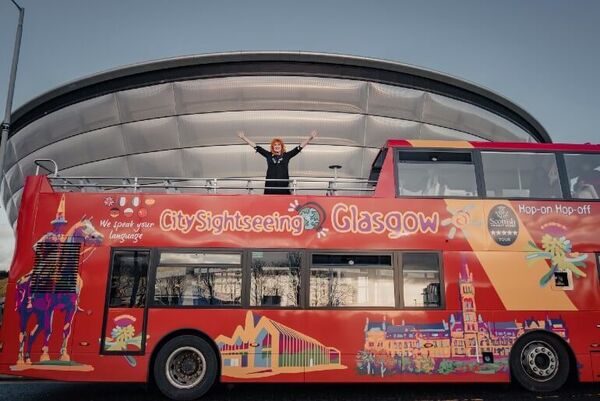 The singer Eddi Reader stands on the top deck of the open top city sightseeing bus with her arms in the air. The bus is parked in front of the silver modern OVO Hydro.