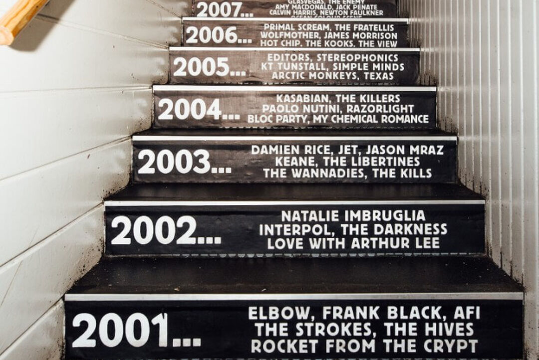 A staircase leading up. Each stair has writing on it including a year and a list of bands that performed in the venue that year.