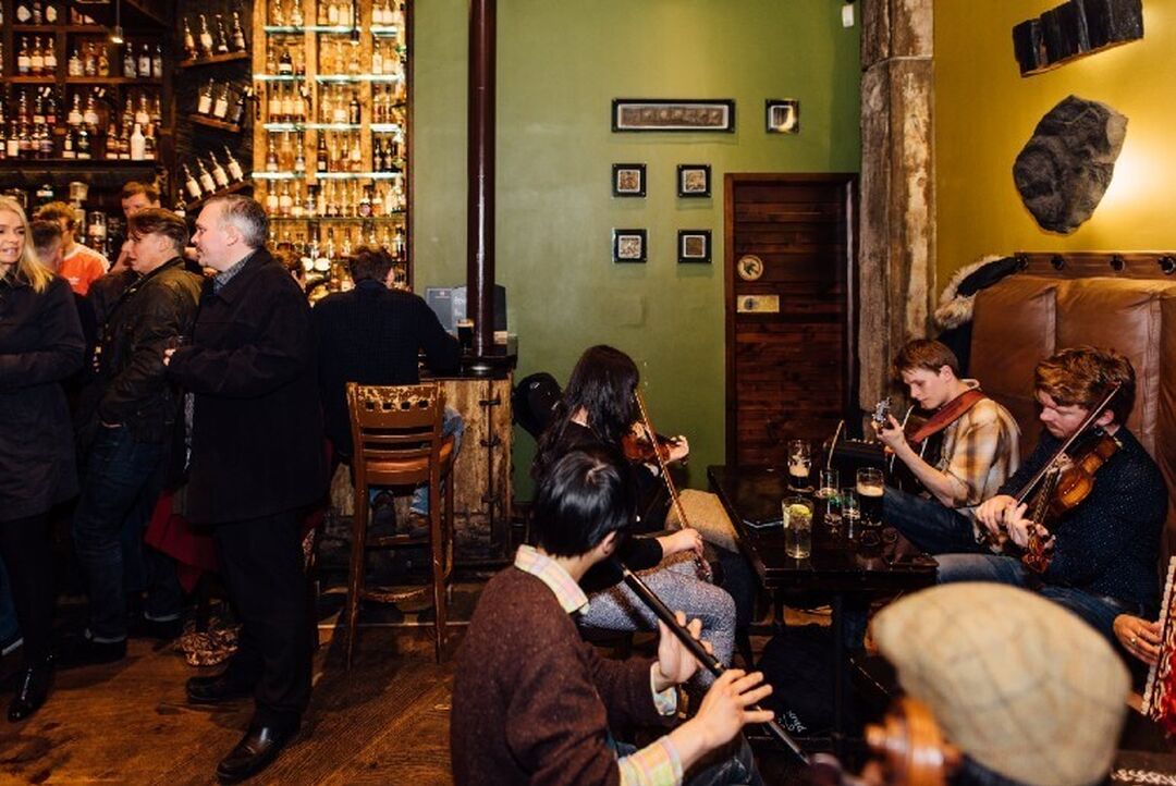 Traditional musicians sit around a table in a pub playing guitars and violins. People stand at the bar area which has a large display of whisky.