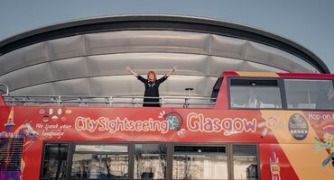 Eddi Reader stands on top of the open top city sightseeing tour bus with her arms in the air. The bus is stopped in front of the silver rounded OVO Hydro venue.