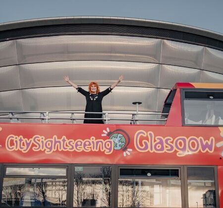 Eddi Reader stands on top of the open top city sightseeing tour bus with her arms in the air. The bus is stopped in front of the silver rounded OVO Hydro venue.