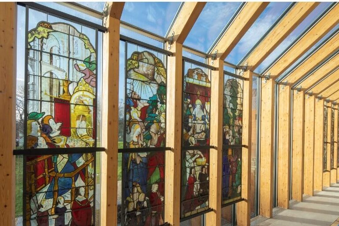 A glass wall and ceiling is lined with a series of wooden beams. 4 of the glass panels have full length stained glass designs.