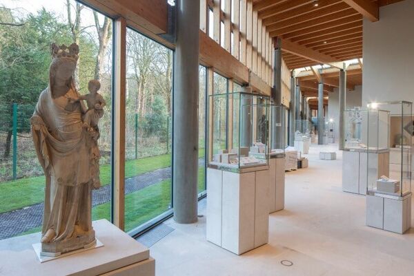 A statue of a person holding a baby is one of the exhibits on a plinth set with a museum. The building wall is made of glass and wood panels and looks out outside onto trees.