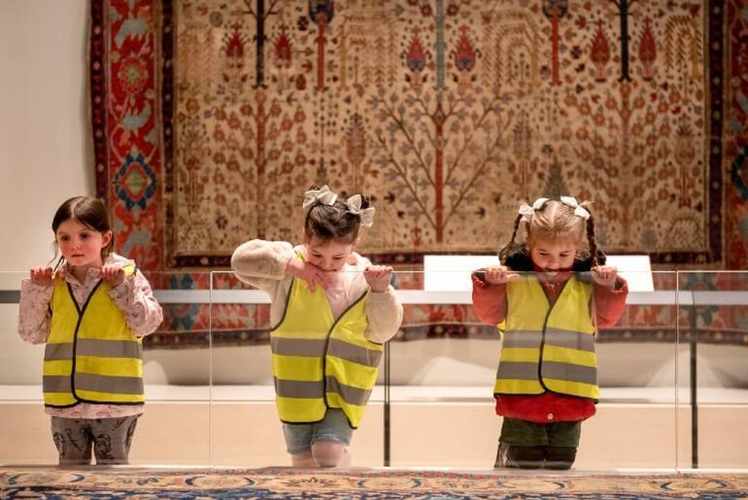 3 children wearing high vis vests lean over a banister looking down. A large rug is displayed on the wall behind them.