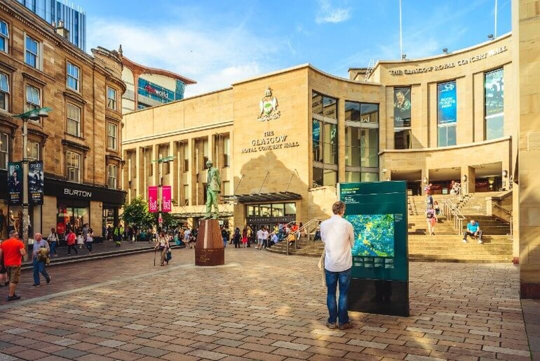 A pedestrianised street leads towards a large building with the sign Glasgow Royal Concert Hall. People walk around and 1 person views a city map.