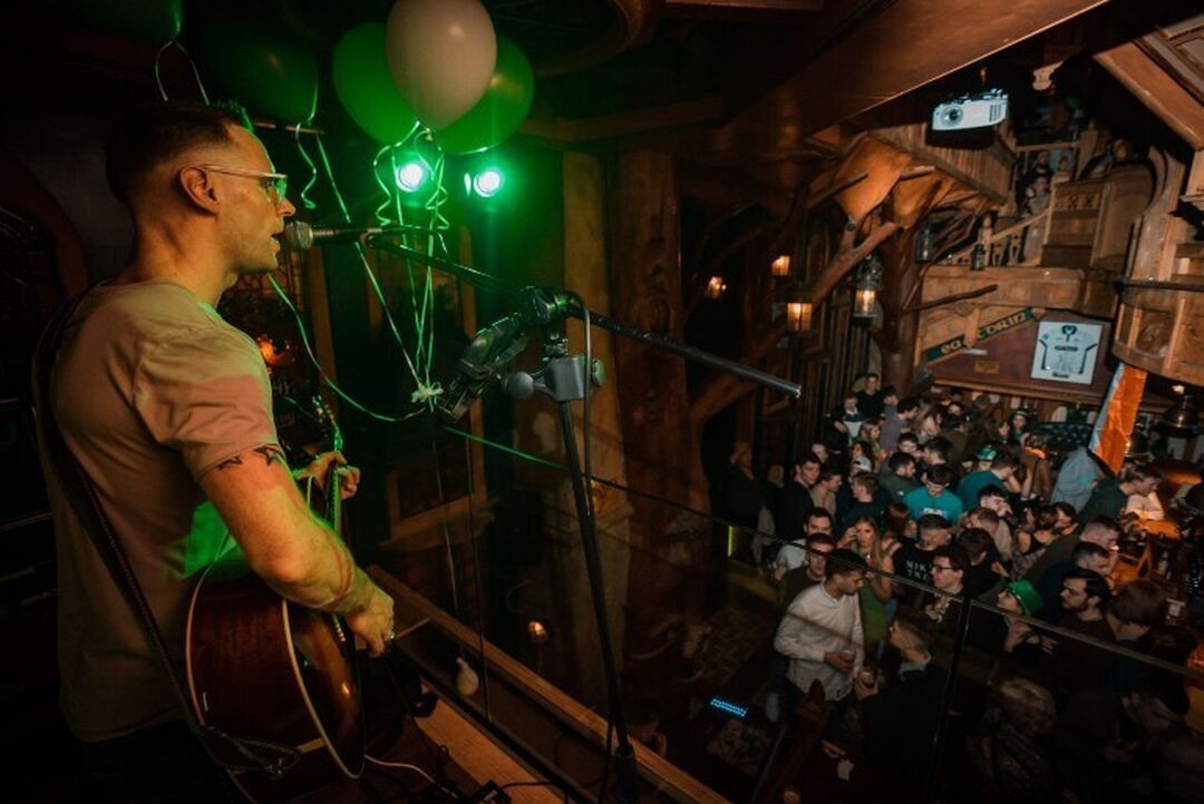 A person playing guitar and singing on a mezzanine level in a wood panelled bar. People are socialising in the bar space below.