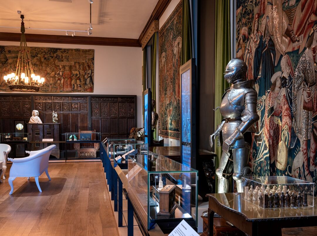 A gallery room full of objects, tapestries, digital screens and a suit of armour.