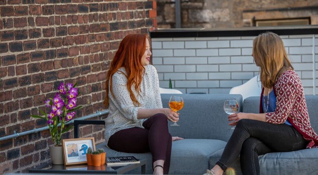 Two people sitting on a sofa talking to each other as they enjoy a drink.