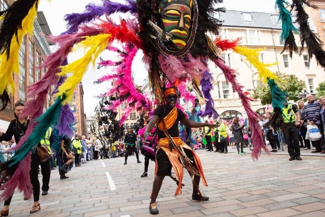 A person with huge colourful feathers and mask leads a carnival parade down a street.