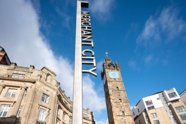 A vertical metal sign reads 'Merchant City'. Behind the sign is a stone tower with clock face and blond sandstone building.