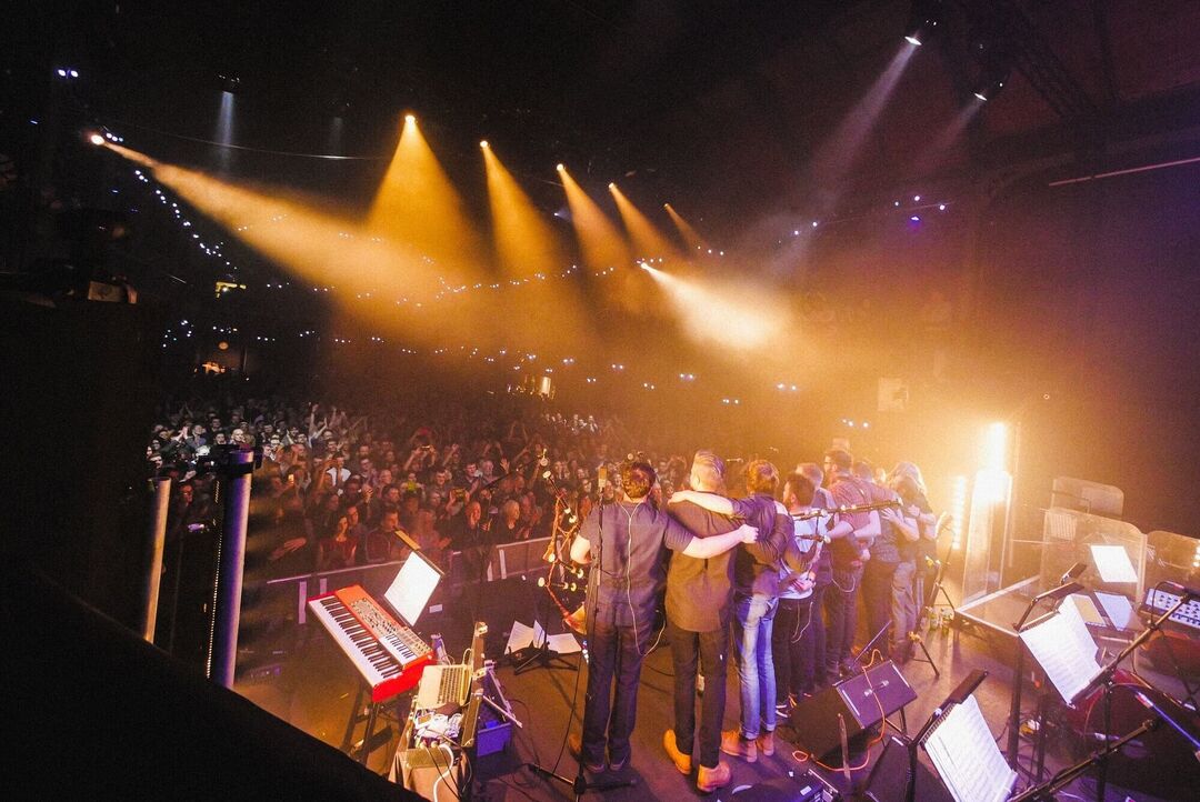 A view from behind and to the left of a group of people on stage at the end of a performance. There is a big crowd clapping.