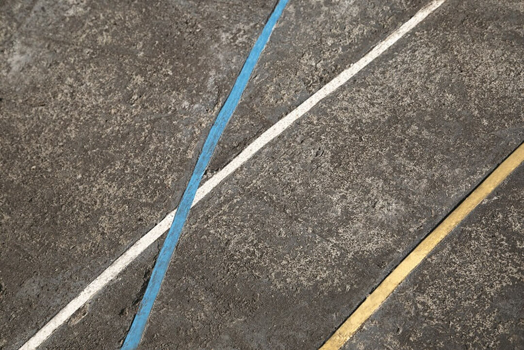 A close-up view of concrete ground with three lines. There is a yellow line and a blue and white line which intersect.