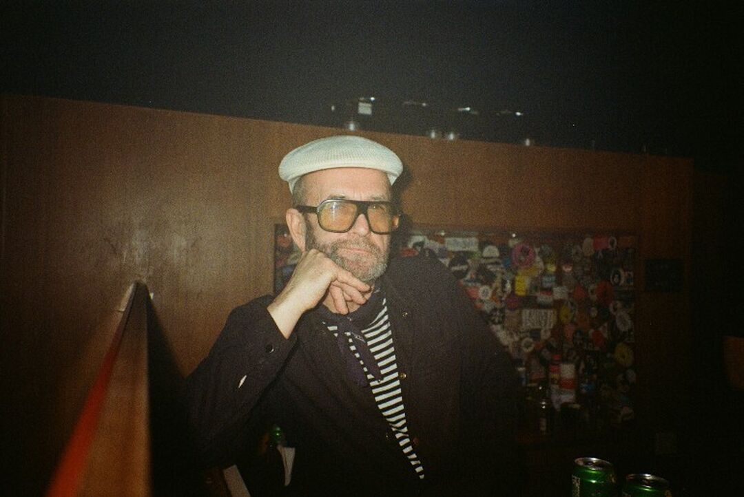 A man leaning on his elbow in a dimly lit room. He wears a white bunnet, square style glasses and a stripy t-shirt under a dark jacket.